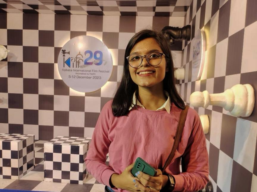 Woman in pink sweater holding a phone in front of a checkered wall with 'IFFI' logo